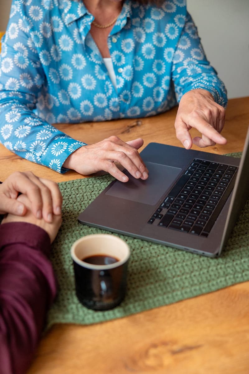 Handen van twee personen bij een laptop; één wijst naar het touchpad, kopje koffie op tafel