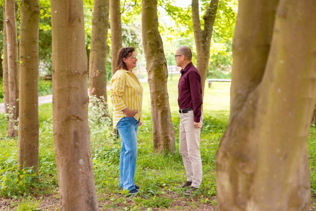Twee volwassenen staan tegenover elkaar en praten tussen bomen in een zonnig park