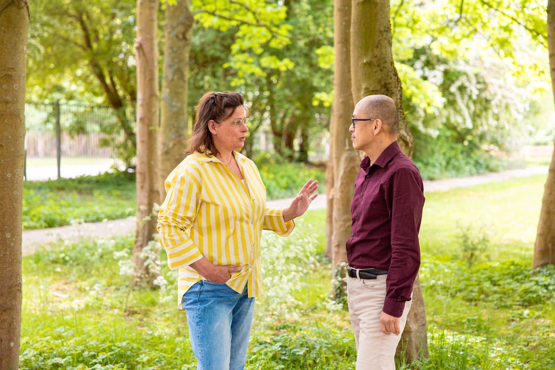 Twee volwassenen praten in een groene parkomgeving tussen bomen; vrouw in geel-witte blouse, man in bordeaux overhemd.