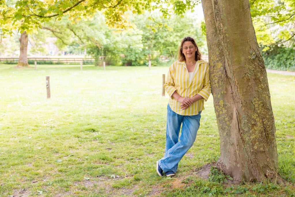 Vrouw leunend tegen een boom in een zonnig park, gele gestreepte blouse en spijkerbroek