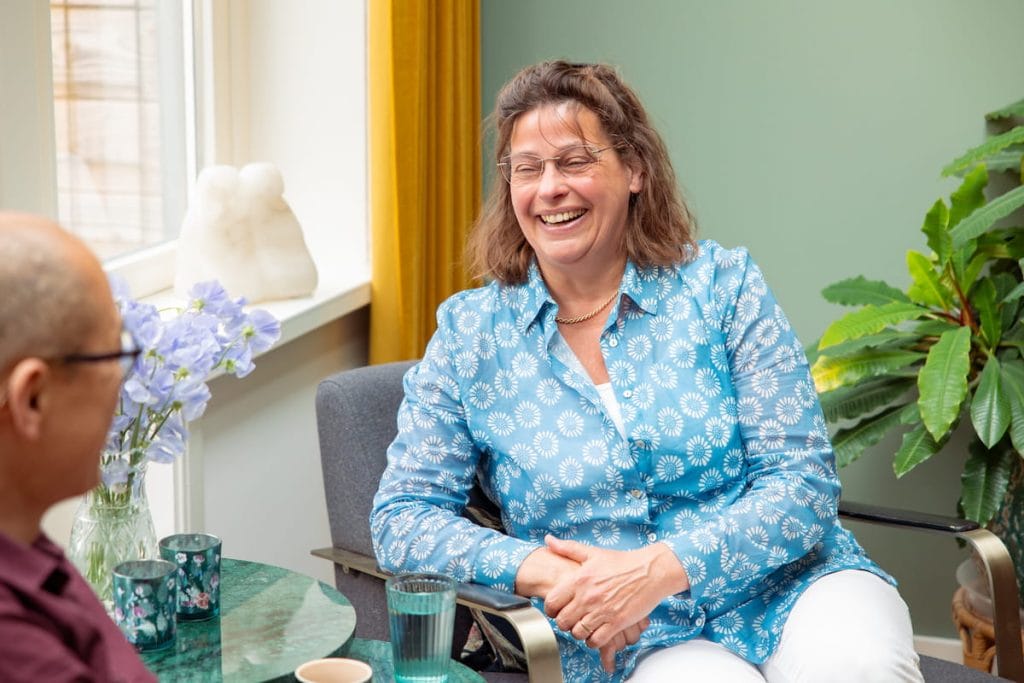 Vrolijke vrouw in blauw overhemd lacht tijdens een ontspannen gesprek in een kantoor van Samen Minder Zorgen