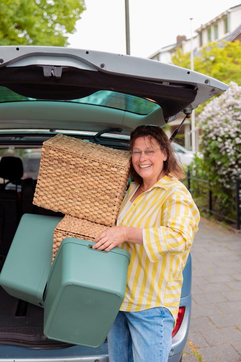 Vrouw lacht terwijl ze gevlochten manden en groene opbergboxen in de kofferbak van een auto laadt