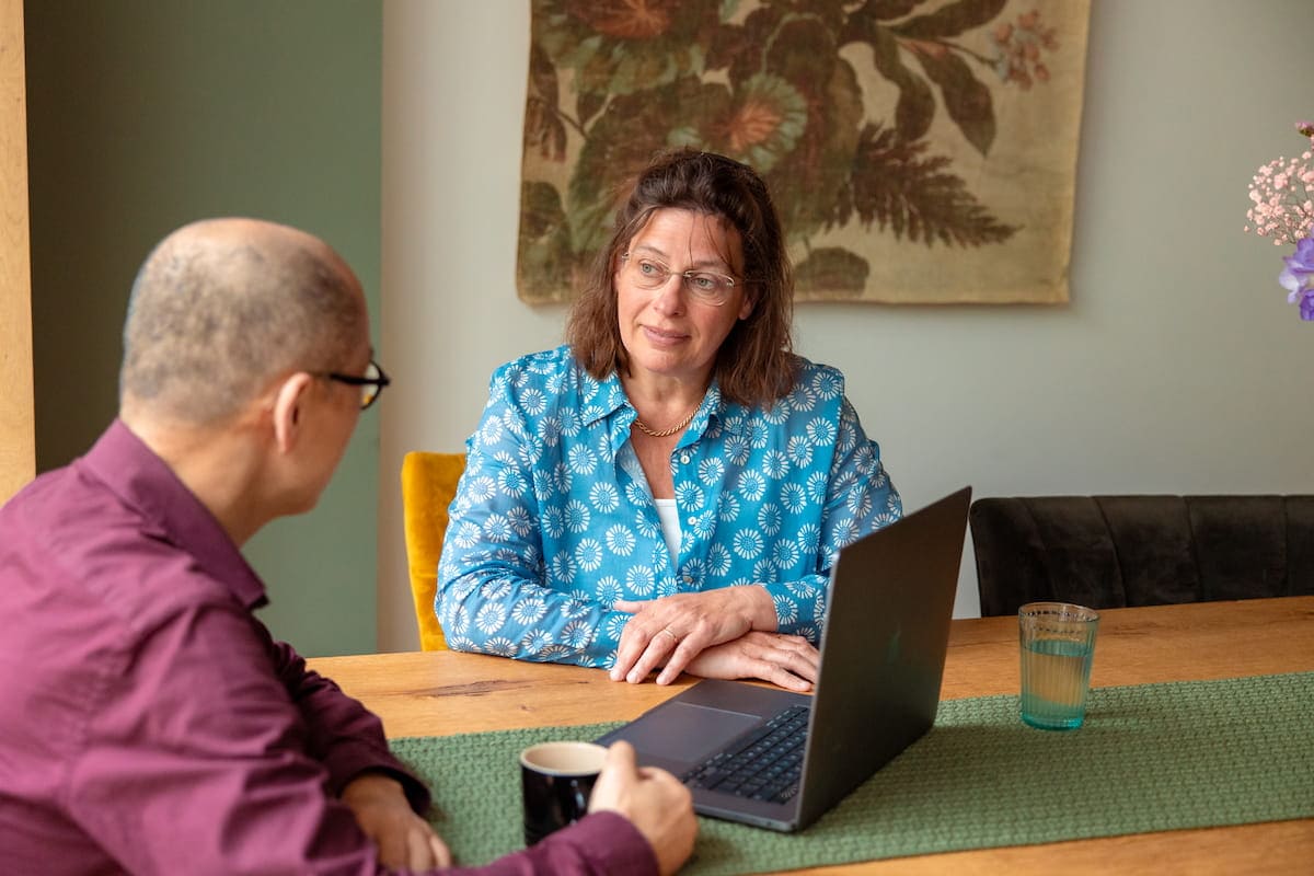 Twee mensen in gesprek aan een houten tafel tijdens een adviesgesprek, laptop, koffiekop en glas water zichtbaar