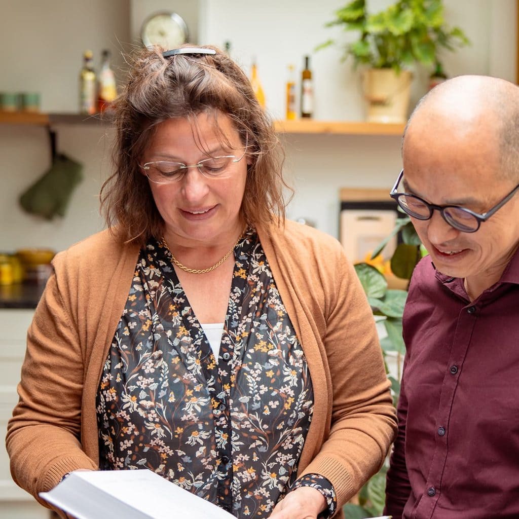 Twee volwassenen lezen samen documenten in een huiselijke keuken, vrouw met bloemendessin en man met bril