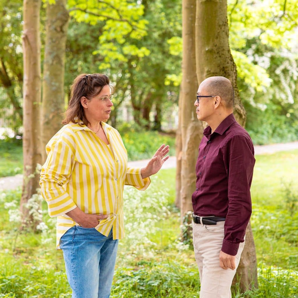 Twee volwassenen in gesprek in een groen park, vrouw met geel-wit gestreepte blouse en man met donkerrood overhemd
