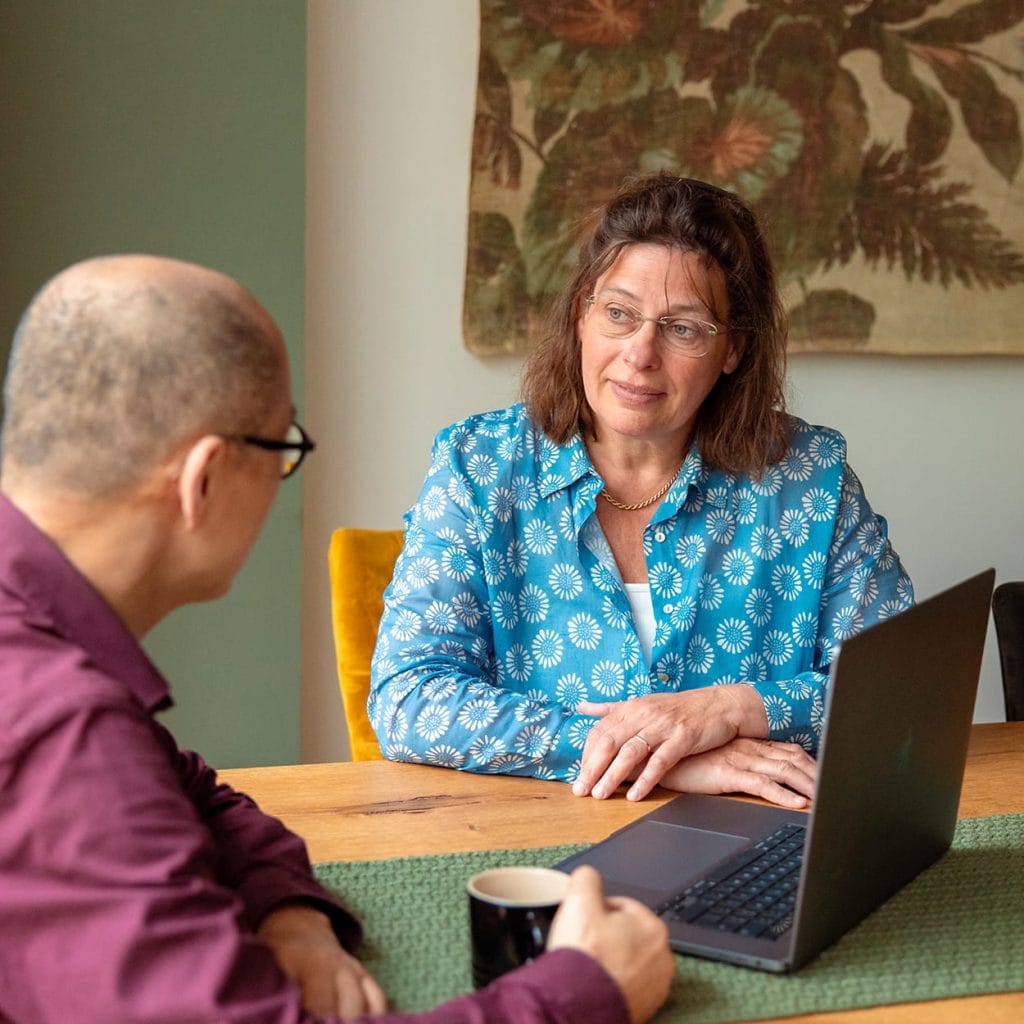 Twee mensen in gesprek aan tafel met laptop en koffie; vrouw in blauw overhemd luistert aandachtig