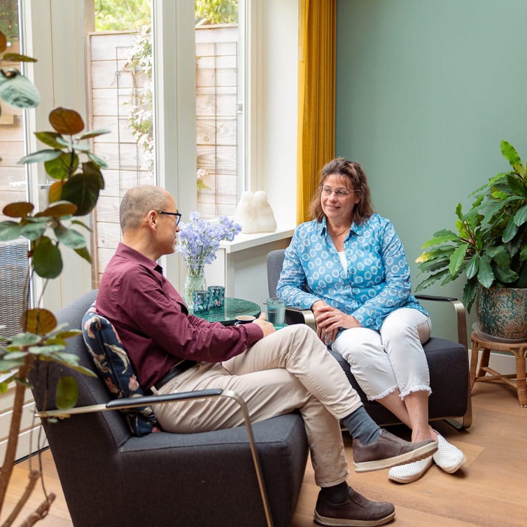 Twee mensen in gesprek in een lichte huiselijke zithoek met planten en groot raam