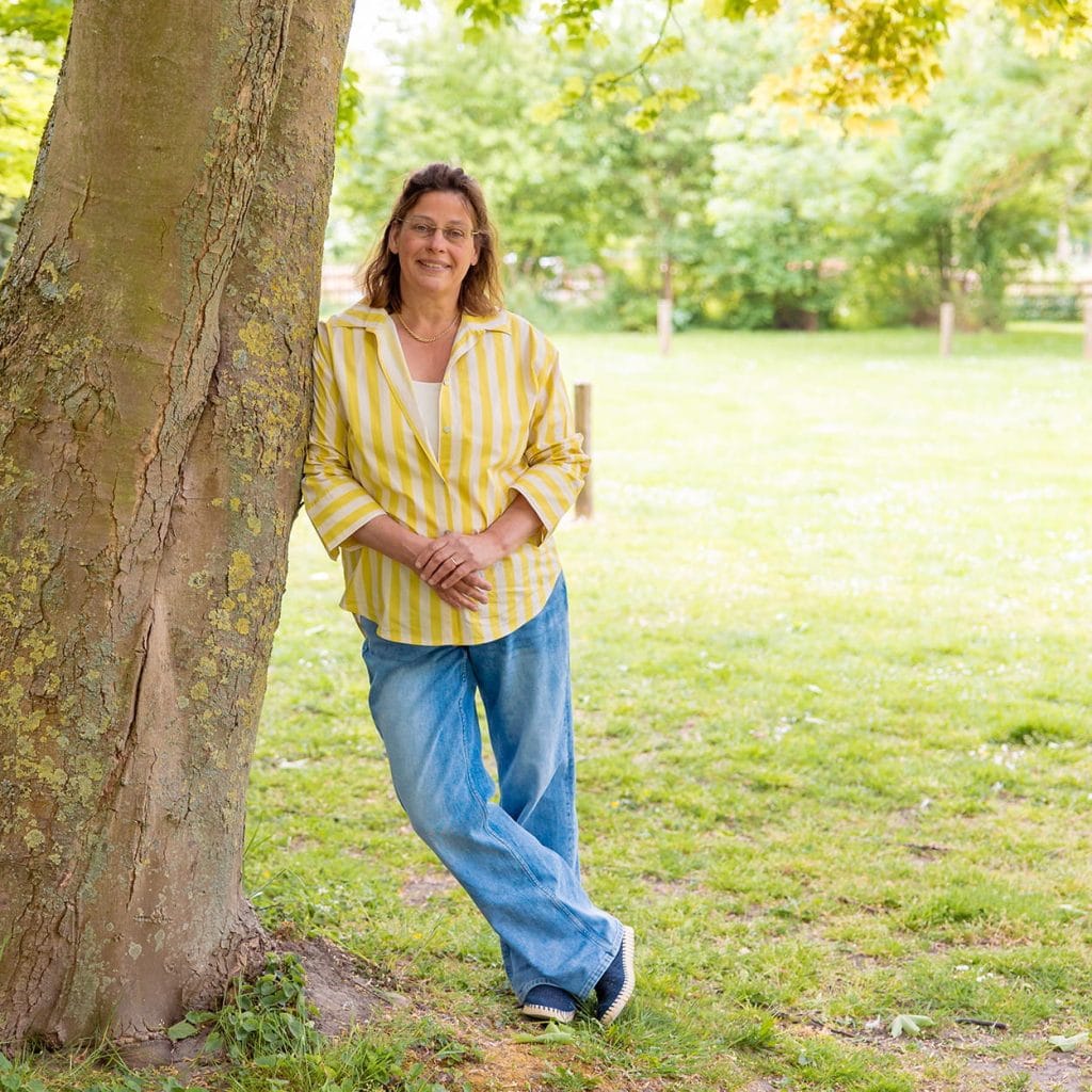 Vrouw leunt tegen boom in een groen park, draagt gele gestreepte blouse en spijkerbroek, glimlacht