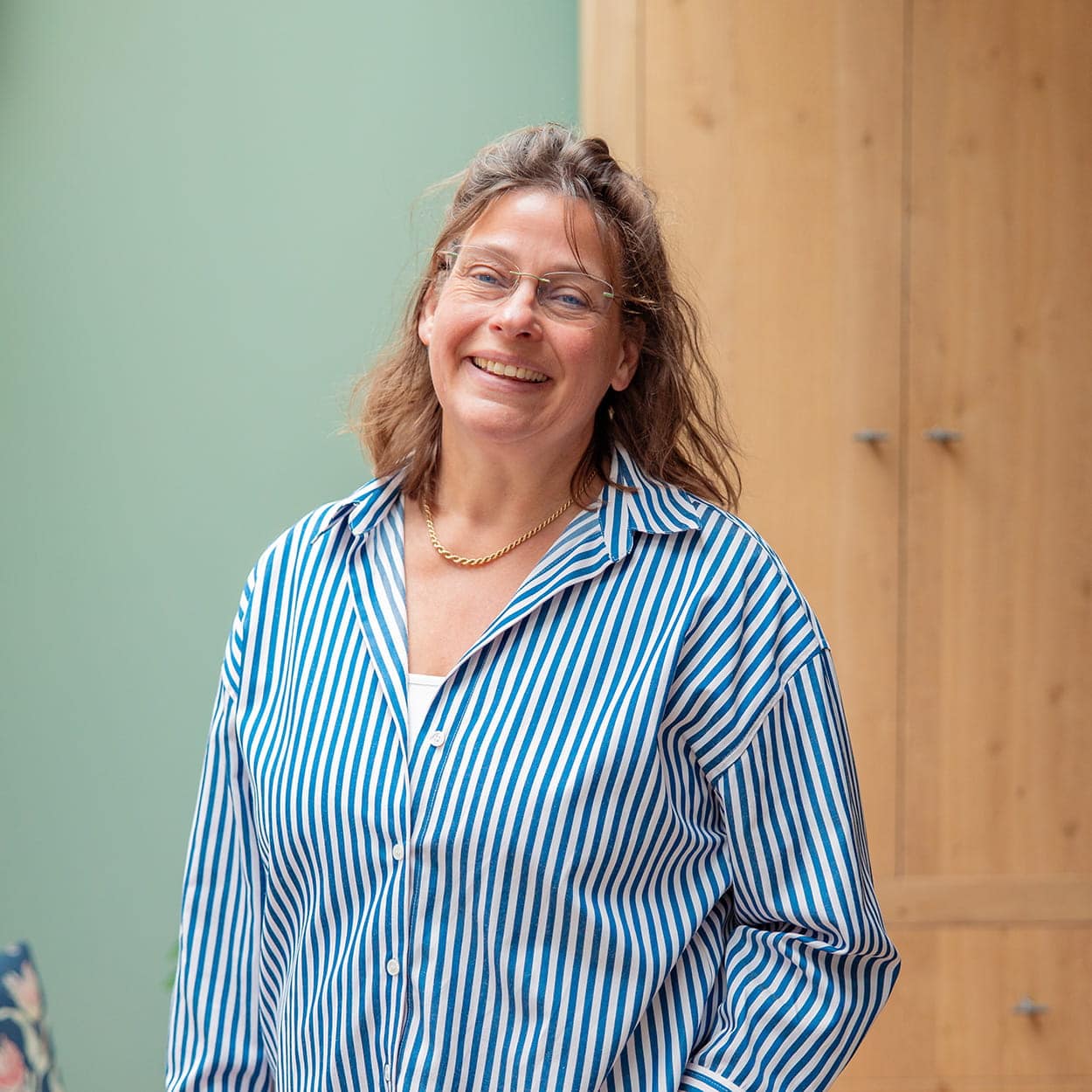 Vrolijke vrouw met gestreept hemd en bril, staand voor groene muur en houten kast